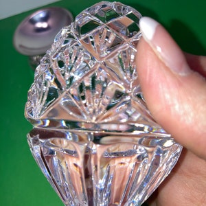 May include: A clear, faceted crystal trinket box with a lid, held by a hand. The box has a diamond pattern and a scalloped edge. A silver-colored object is visible in the background.