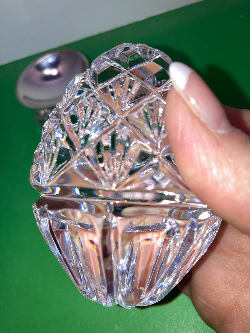 May include: A clear, faceted crystal trinket box with a lid, held by a hand. The box has a diamond pattern and a scalloped edge. A silver-colored object is visible in the background.