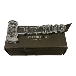 May include: A clear crystal gavel featuring intricate cross-hatch and diamond-cut patterns on the head and handle. The gavel rests horizontally on a dark rectangular box with the white text WATERFORD CRYSTAL printed on the top surface.