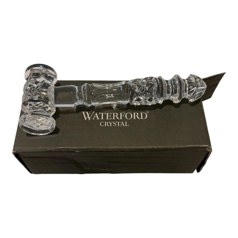 May include: A clear crystal gavel featuring intricate cross-hatch and diamond-cut patterns on the head and handle. The gavel rests horizontally on a dark rectangular box with the white text WATERFORD CRYSTAL printed on the top surface.