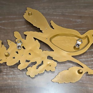 Pode incluir: Decora&ccedil;&atilde;o de parede dourada em forma de p&aacute;ssaro pousado em um galho com folhas. A parte de tr&aacute;s do p&aacute;ssaro e do galho tem dois cabides de metal para montagem. O p&aacute;ssaro est&aacute; voltado para a direita.