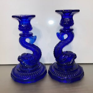 May include: A pair of cobalt blue glass candle holders shaped like dolphins. The candle holders have a detailed design and a scalloped base.
