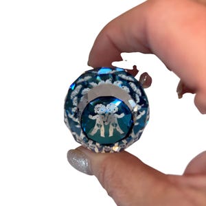 Könnte beinhalten: Ein facettierter, blau-klarer Kristallschmuck mit einem Zwillinge-Symbol im Inneren. Der Schmuck wird zwischen Fingern vor weißem Hintergrund gehalten. Das Zwillinge-Symbol zeigt zwei Strichmännchen, die sich an den Händen halten.