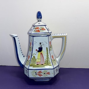 May include: White ceramic teapot with a blue and gold handle. The teapot is decorated with a folk art style design featuring a woman in a red and yellow dress, flowers, and birds.