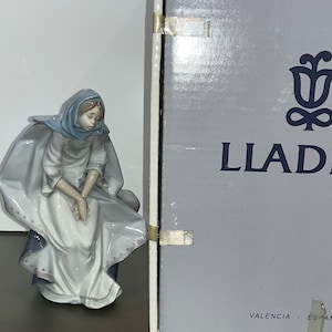 Può includere: Una statuetta in porcellana di una donna vestita con un abito blu e bianco e uno scialle blu. La statuetta è seduta su una sedia e ha un'espressione triste sul viso. La statuetta è realizzata da Lladró, un'azienda spagnola nota per le sue statuette in porcellana di alta qualità.