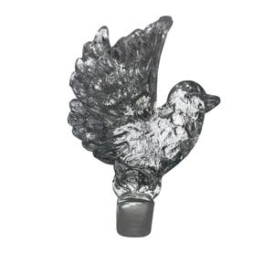 Puede incluir: Un tapón de botella de vidrio transparente con forma de pájaro con las alas extendidas. El pájaro tiene plumas detalladas y una base cilíndrica gris. El tapón está diseñado para encajar en una botella.