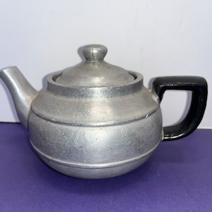 May include: A silver teapot with a black handle. The teapot has a rounded body and a lid with a knob on top.
