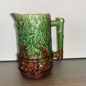 Puede incluir: Jarra de cerámica verde y marrón con diseño de hojas. La jarra tiene un asa que se asemeja a una rama de árbol. La base de la jarra tiene un diseño floral marrón. La jarra es probablemente vintage o antigua.