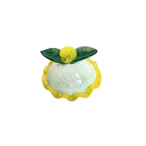 May include: A small glass figurine of a flower with a yellow centre and green leaves. The flower is sitting on a white base with a yellow rim.
