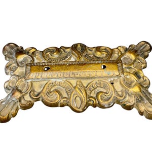 May include: Ornate, antique gold-colored metal decorative piece with a rectangular center and scrolling floral designs. The piece has four feet and two holes in the center. The metal has a distressed finish.