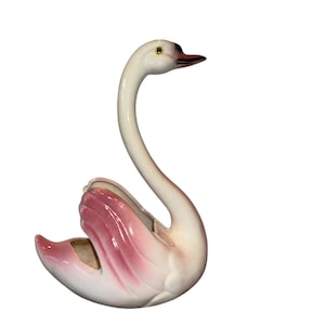 May include: A ceramic swan ornament with a long, curved neck and a graceful pose. The swan is mainly white with pink accents on its wings. The beak is dark, and the eyes are yellow.
