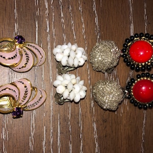 May include: Six pairs of vintage clip-on earrings. Two pairs are pink with gold accents and a purple stone. Two pairs are white and made of small shells. Two pairs are gold with black beads and a red stone.