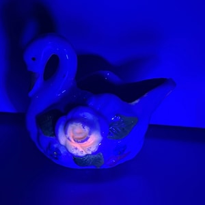 May include: A blue ceramic swan planter with a pink rose and green leaves. The planter is illuminated with a blue light.