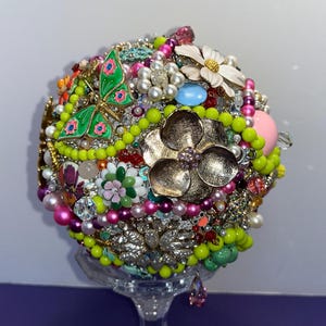 May include: A round, colorful bouquet made of various brooches, beads, and rhinestones. The bouquet features a green butterfly brooch, a gold flower brooch, and a white flower brooch. The bouquet is displayed on a clear glass stand.