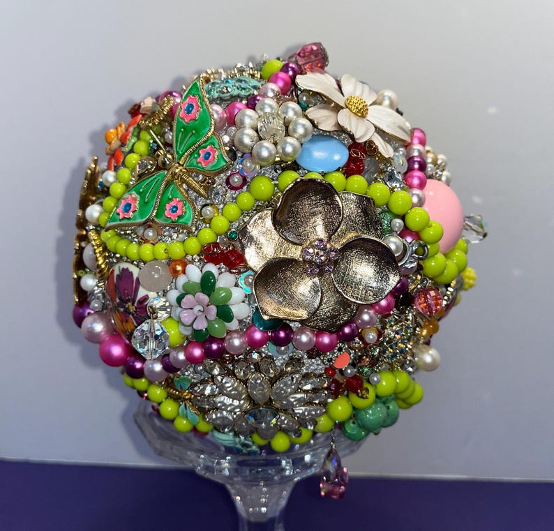 May include: A round, colorful bouquet made of various brooches, beads, and rhinestones. The bouquet features a green butterfly brooch, a gold flower brooch, and a white flower brooch. The bouquet is displayed on a clear glass stand.