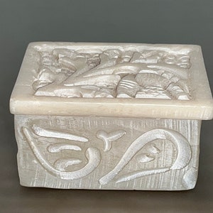 May include: A small, white, rectangular box with intricate carved floral designs on the top and sides. The box is made of a smooth, polished material.