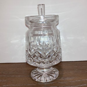 Op de afbeelding: Een glazen pot met een deksel en een voet. De pot heeft een geslepen kristallen ontwerp.