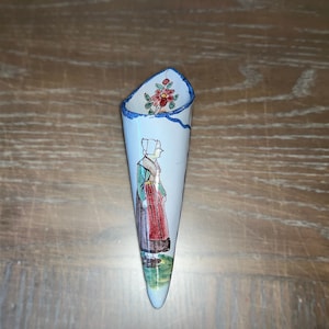 Puede incluir: Un portaobjetos de pared de cerámica blanca en forma de cono con un borde azul y una escena pintada de una mujer con un vestido verde y una falda roja. La mujer está de pie frente a una flor roja.