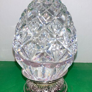 May include: A clear, cut-glass decorative object with a diamond pattern, resting on a silver-colored base with an ornate design. The object has a rounded, egg-like shape.