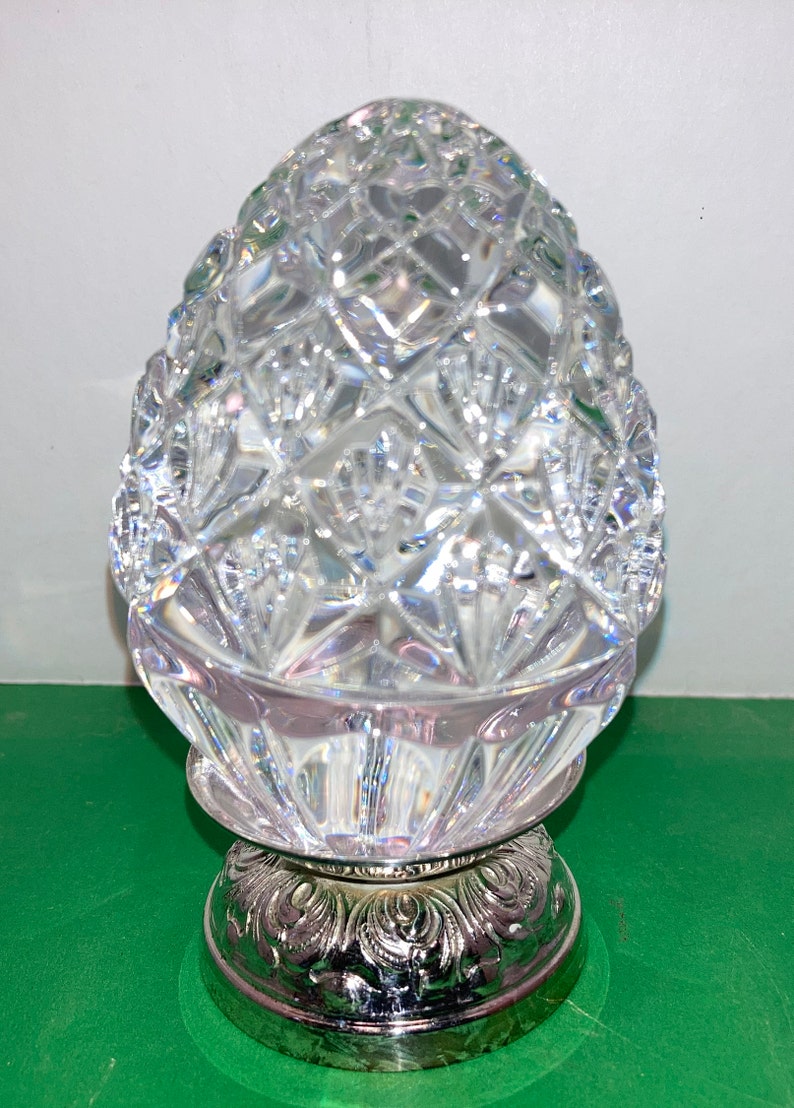 May include: A clear, cut-glass decorative object with a diamond pattern, resting on a silver-colored base with an ornate design. The object has a rounded, egg-like shape.