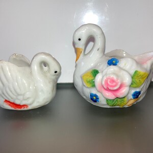 May include: Two white porcelain swan figurines. One swan has a pink and white rose with green leaves and blue flowers painted on its body. The other swan has a red stripe painted on its body.
