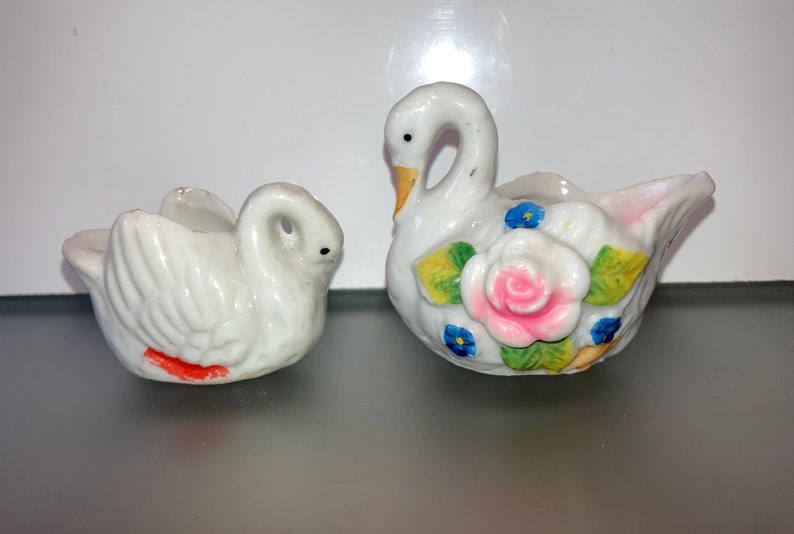 May include: Two white porcelain swan figurines. One swan has a pink and white rose with green leaves and blue flowers painted on its body. The other swan has a red stripe painted on its body.