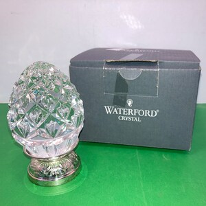 May include: A clear crystal pineapple-shaped decorative object with a silver-colored base. The pineapple has a textured, diamond-cut pattern. A gray box with the words "WATERFORD CRYSTAL" is behind the object.