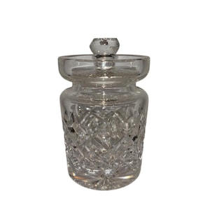 Könnte beinhalten: Ein klarer, geschliffener Glasbehälter mit Deckel. Der Behälter hat ein Rautenmuster und einen kleinen Knopf auf dem Deckel. Der Behälter wird wahrscheinlich zur Aufbewahrung kleiner Gegenstände oder als Dekorationsstück verwendet.