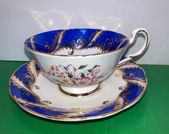 May include: A vintage teacup and saucer set. The teacup has a white interior with a floral design and a cobalt blue exterior rim with gold filigree. The saucer mirrors the teacup's design, featuring a cobalt blue border and gold accents.