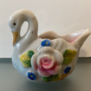 May include: A white porcelain swan figurine with a pink and white rose and blue flowers. The swan has a yellow beak and is shaped like a small dish.