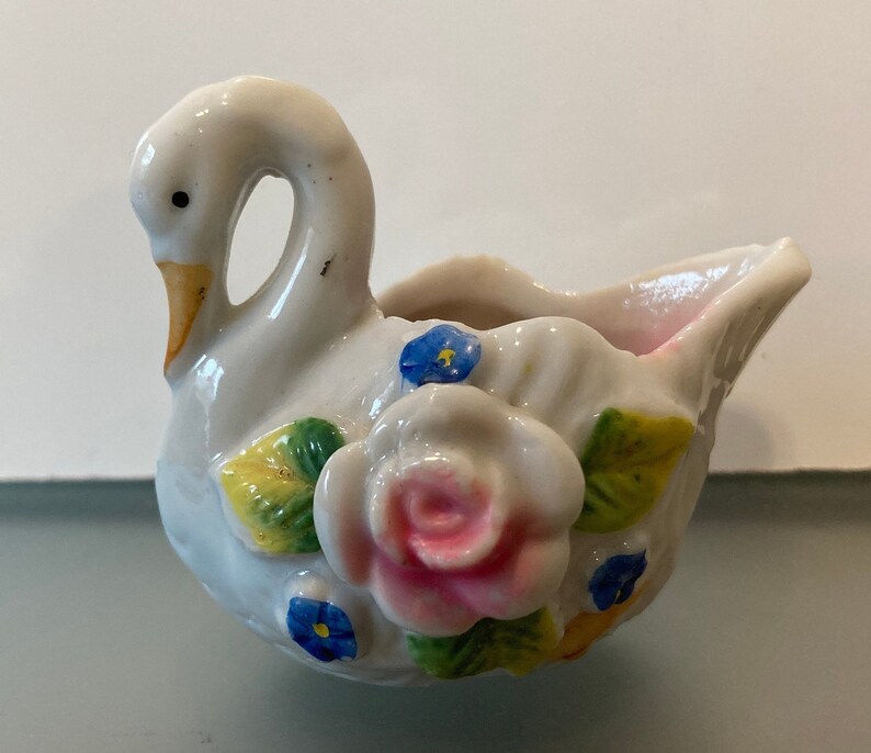 May include: A white porcelain swan figurine with a pink and white rose and blue flowers. The swan has a yellow beak and is shaped like a small dish.