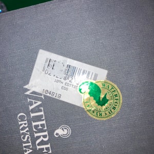 May include: Close-up of a dark gray box with a white sticker and a gold and green sticker. The white sticker has a barcode and text that reads "18th Edition Egg". The gold sticker says "Waterford Crystal".