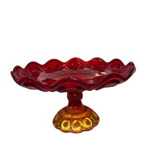 May include: A ruby red glass cake stand with a scalloped edge. The base is a golden amber color with a starburst design. The stand is a decorative piece for displaying desserts or other items.
