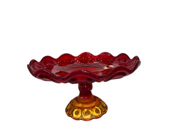 Puede incluir: Un soporte para pasteles de vidrio rojo rubí con borde festoneado. La base es de color ámbar dorado con un diseño de estrella. El soporte es una pieza decorativa para exhibir postres u otros artículos.