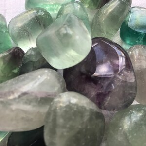 May include: A collection of tumbled fluorite stones in shades of green, purple, and white. The stones are smooth and polished, with a variety of shapes and sizes.