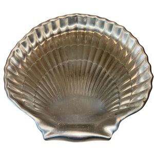 May include: A silver-colored, scallop-shaped dish with a textured surface. The dish has a wide, shallow bowl and a scalloped edge, resembling a seashell. The dish is made of metal and is likely used for serving or display.