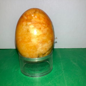 May include: A smooth, yellow and orange marble egg on a clear plastic stand. The egg has a swirling pattern of colour.