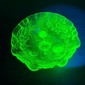 Może przedstawiać: Okrągła szklana miska z falistym brzegiem, świecąca jaskrawą neonową zielenią w świetle ultrafioletowym. Wewnętrzna powierzchnia posiada szczegółowy, tłoczony wzór kwiatowy z różami i liśćmi oraz teksturowany motyw wachlarza na krawędzi.