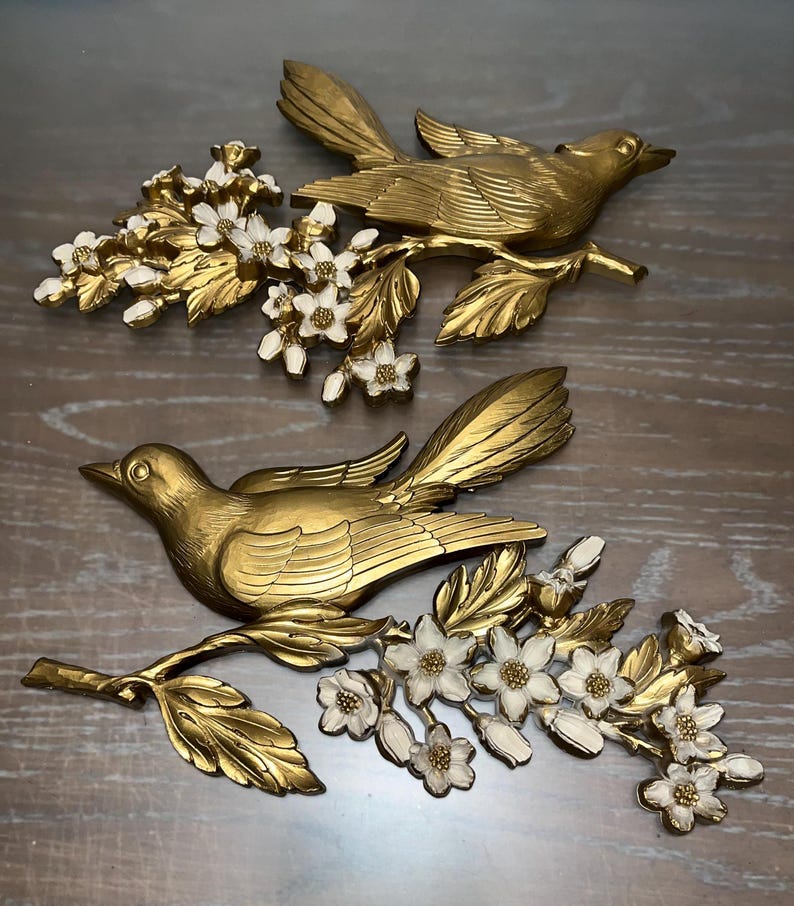 Pode incluir: Duas esculturas de parede de p&aacute;ssaros dourados com asas abertas, pousadas em galhos com flores brancas e douradas. Os p&aacute;ssaros s&atilde;o detalhados com penas e bicos texturizados, criando uma est&eacute;tica vintage.
