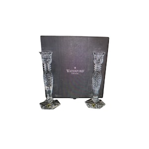 May include: Two clear crystal candlesticks with intricate cut glass patterns, flared tops, and hexagonal bases. Each candlestick features a small green and gold sticker. Behind them sits a dark grey box with the text WATERFORD CRYSTAL.