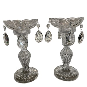 Op de afbeelding: Twee heldere kristallen kandelaars met sierlijke details. Elke houder heeft een geschulpte bovenkant, een gedraaide steel en een ronde basis. Kristallen druppelhangers hangen van de bovenkant.