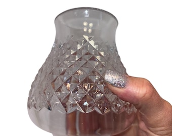 May include: A clear, diamond-cut glass vase with a rounded base and flared top. The vase is held by a hand with glittery nail polish. The glass reflects light, creating a sparkling effect. The vase is empty.