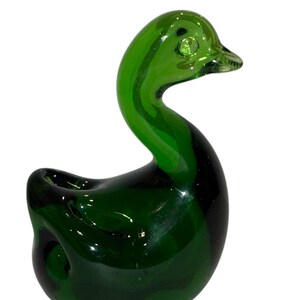 May include: A green glass duck figurine. The duck has a rounded body and a long neck. The duck's eyes are made of clear glass.