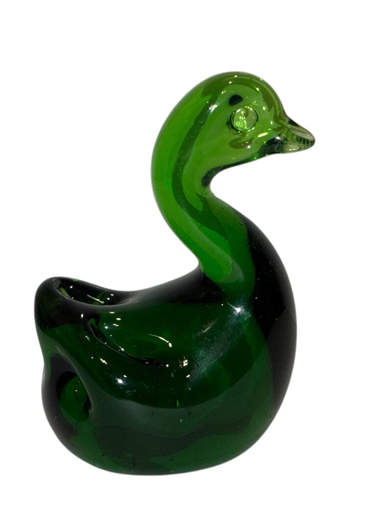 May include: A green glass duck figurine. The duck has a rounded body and a long neck. The duck's eyes are made of clear glass.