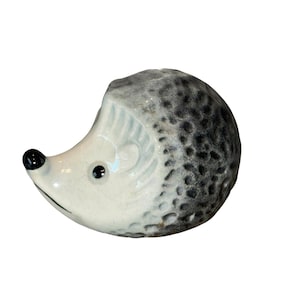 May include: A ceramic hedgehog figurine with a white face, gray and black speckled body, and black eyes and nose. The figurine is a decorative item, likely for home decor.