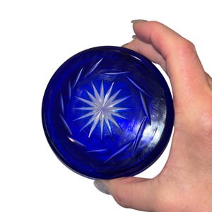 Może przedstawiać: Okrągła, kobaltowa szklana pokrywka z wzorem rozety w środku. Pokrywka trzymana jest w dłoni na białym tle. Rozeta jest biała, a reszta pokrywki ma głęboki niebieski kolor.