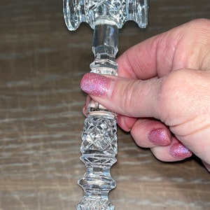 May include: A clear, cut-glass cabinet handle with a decorative, symmetrical design. The handle features a cylindrical stem with diamond-shaped cuts and two rounded ends. The handle is held in a person's hand.