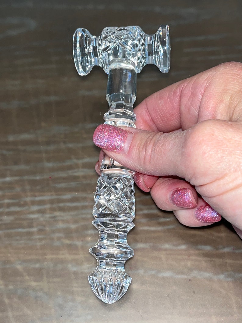 May include: A clear, cut-glass cabinet handle with a decorative, symmetrical design. The handle features a cylindrical stem with diamond-shaped cuts and two rounded ends. The handle is held in a person's hand.