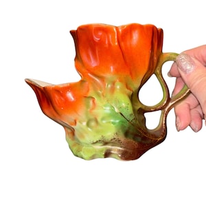 Puede incluir: Una taza de cerámica con forma de tulipán y un asa. La taza presenta un degradado de colores, que va del naranja en la parte superior al verde y marrón en la base. El asa tiene un diseño curvo y la taza tiene una superficie texturizada única.