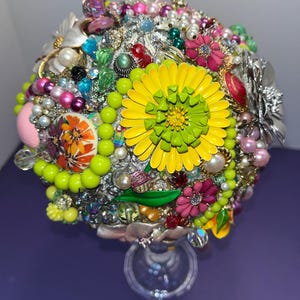 May include: A colorful bouquet made of vintage brooches and jewelry, featuring a large yellow flower with a green center. The bouquet is arranged on a clear glass stand.